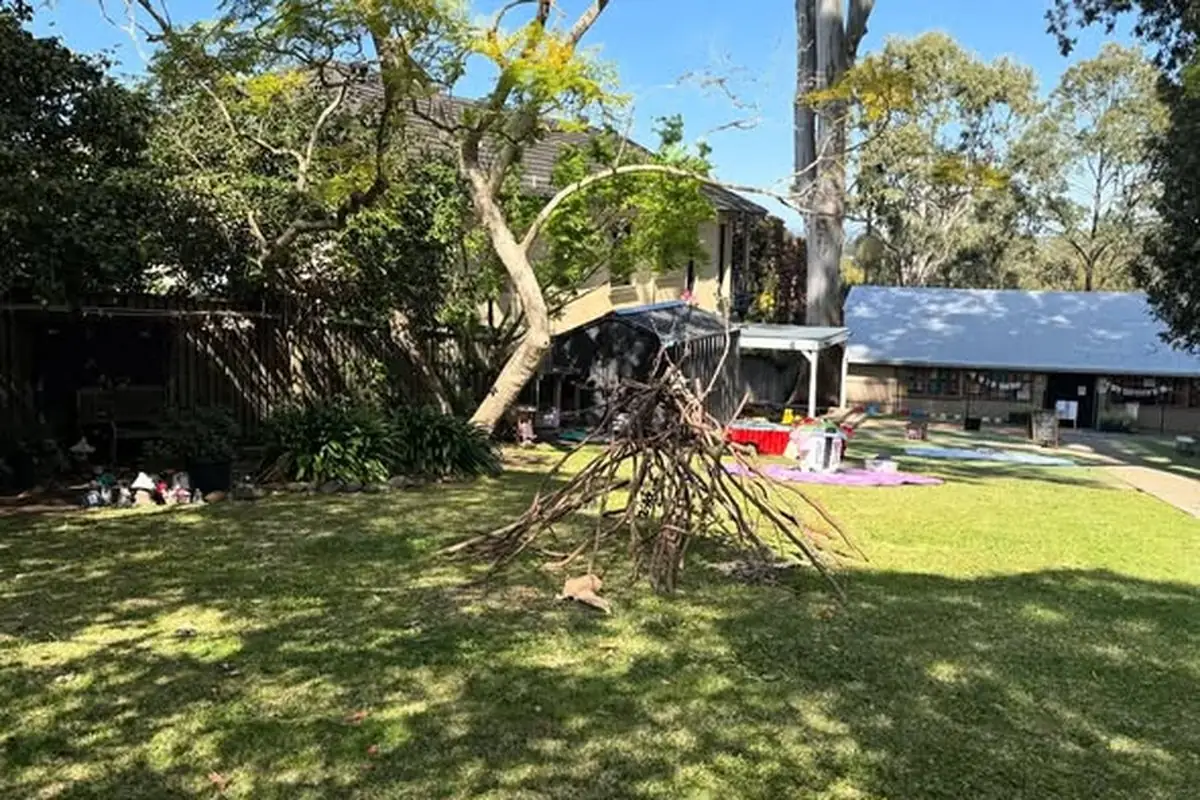 Wide open playground with a teepee-shaped branch structure, shaded by gum trees and dappled sunlight. Children's learning materials set out on rugs in the distance.