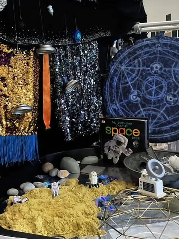 A World Space Week invitation to play: sequin fabrics as galaxies, a starry backdrop, a pop-up space book, and a kinetic sand tray with tiny astronauts exploring craters.